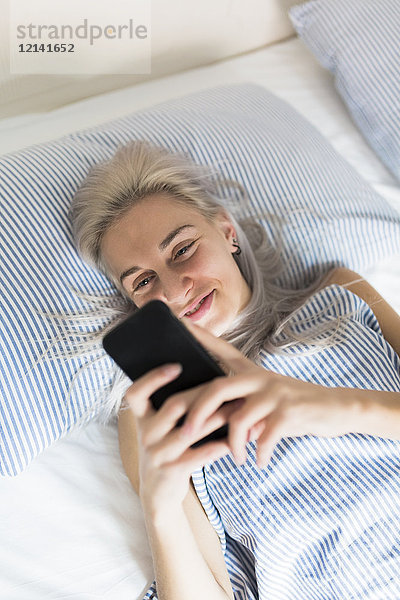 Lächelnde junge Frau  die im Bett liegt und ihr Handy überprüft.