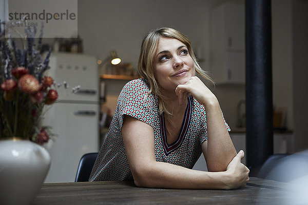 Porträt einer lächelnden Frau  die am Tisch in der Küche sitzt und nachdenkt.