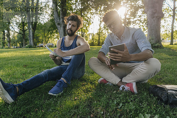 Zwei Freunde sitzen in einem Park mit mobilem Gerät und Papieren