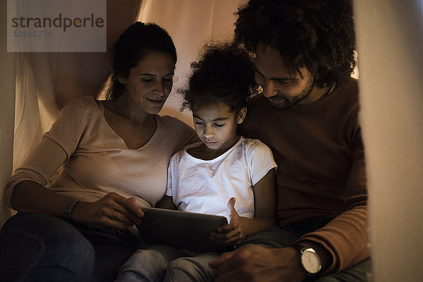Eltern sitzen im Kinderzimmer mit Tochter und schauen auf das digitale Tablett.