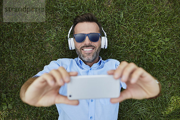Porträt eines lachenden Mannes auf einer Wiese liegend  Selfie mit Smartphone  Draufsicht
