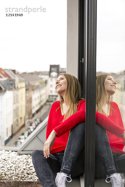 Lächelnde junge Frau sitzt am Fenster in der Stadtwohnung