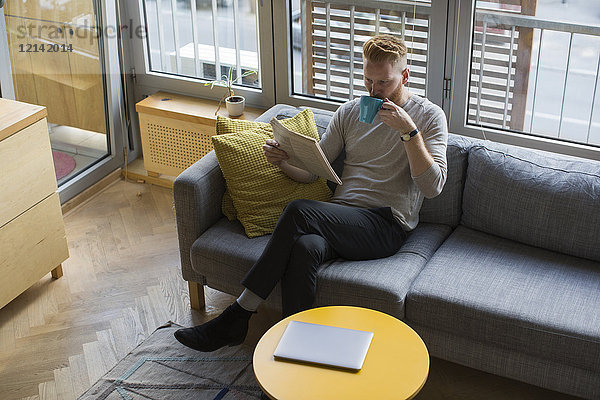 Mann sitzt zu Hause auf der Couch und liest Zeitung und trinkt Kaffee.
