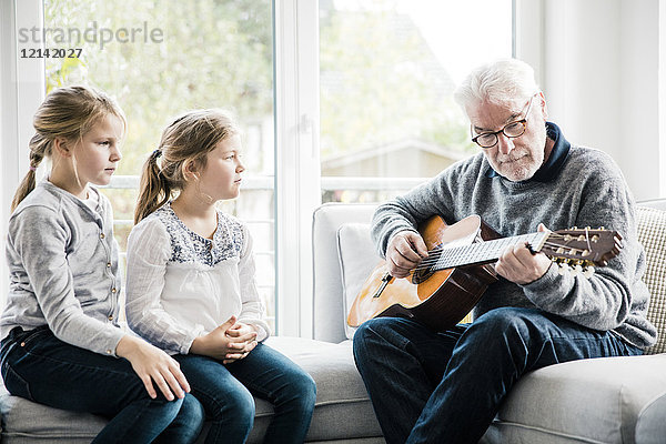 Zwei Mädchen sitzen auf dem Sofa und hören Großvater beim Gitarrespielen zu.