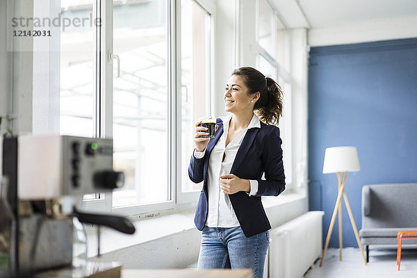 Fröhliche Geschäftsfrau mit einem Glas Kaffee in einem Loft aus dem Fenster schauend