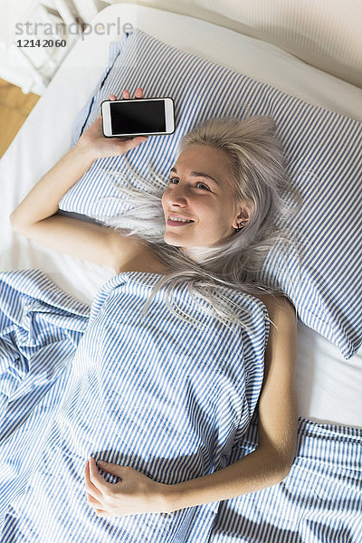Lächelnde junge Frau im Bett liegend mit Handy