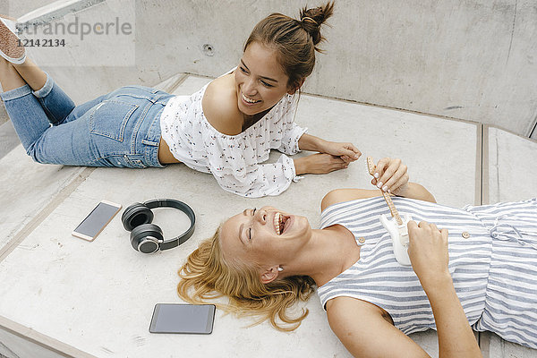 Zwei glückliche junge Frauen  die auf einer Rampe in einem Skatepark liegen und Musik machen.