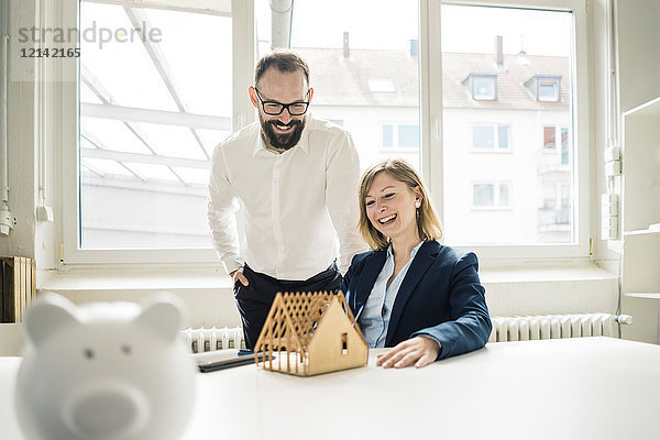 Lachende Frau und Mann mit Hausmodell und Sparschwein im Büro