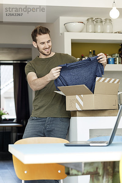 Lächelnder junger Mann zu Hause beim Auspacken des Pakets mit Kleidung