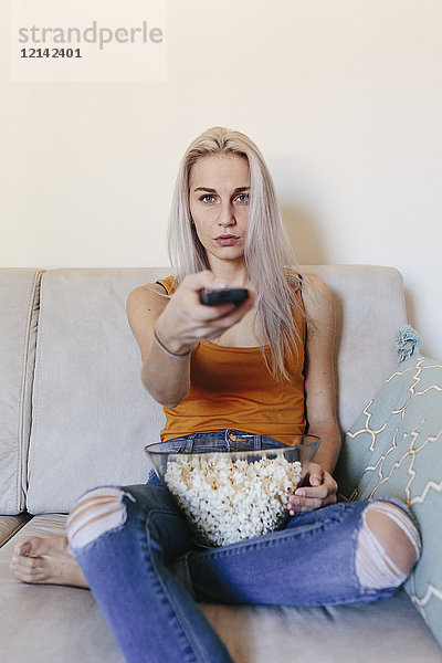 Junge Frau zu Hause auf der Couch sitzend mit Popcorn und Fernbedienung
