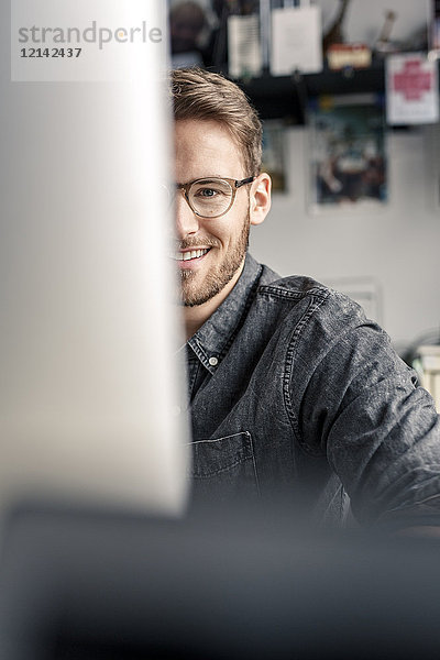 Porträt eines lächelnden jungen Mannes hinter dem Computerbildschirm am Schreibtisch zu Hause