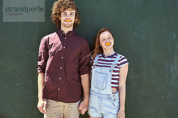 Junges Paar steht vor einer grünen Wand mit Orangenscheiben im Mund.
