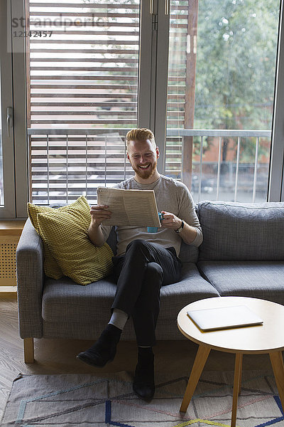 Lächelnder Mann sitzt zu Hause auf der Couch und liest Zeitung.