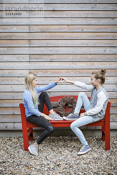 Zwei beste Freunde sitzen auf einer roten Bank vor einer Holzfassade und bilden mit ihren Händen ein Herz.