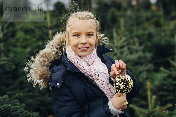 Kleines Mädchen steht vor Tannenbäumen und hält einen schokoladengetauchten Apfel.
