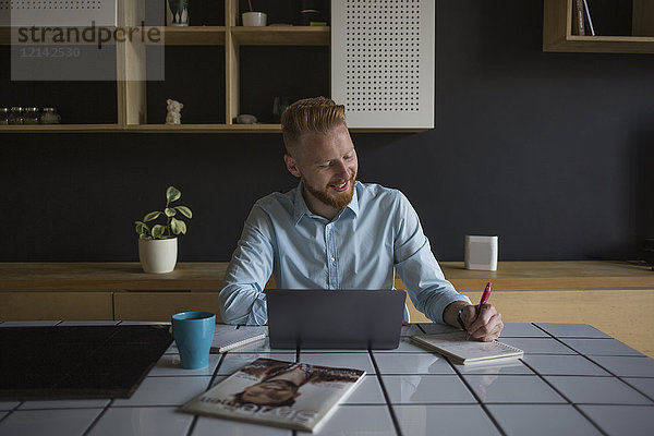 Lächelnder Geschäftsmann mit Laptop und Notebook bei der Arbeit zu Hause