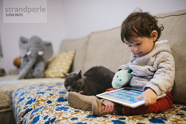 Kleines Mädchen sitzt auf der Couch und schaut sich Videos auf einem Tablett mit einer Katze an.