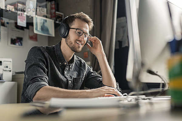 Lächelnder junger Mann mit Kopfhörern arbeitet am Computer zu Hause am Schreibtisch
