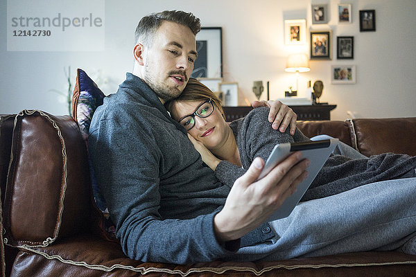 Lächelndes Paar liegt auf der Couch zu Hause und teilt sich das Tablett.