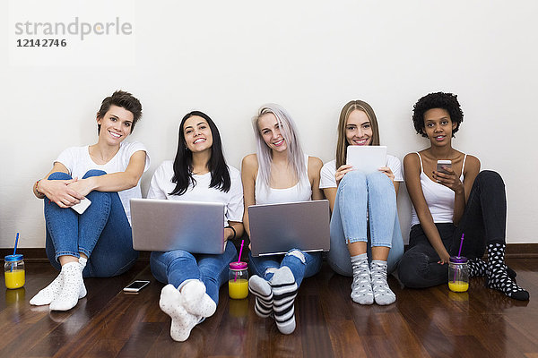 Porträt von lächelnden Freundinnen zu Hause auf dem Boden sitzend mit Technik