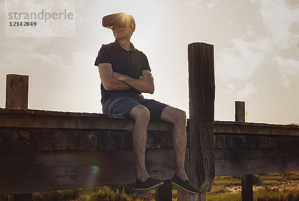Erwachsener Mann mit VR-Brille  der bei Sonnenuntergang auf der Strandpromenade sitzt und die Aussicht genießt.