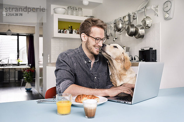 Lächelnder Mann mit Hund mit Laptop in der Küche zu Hause