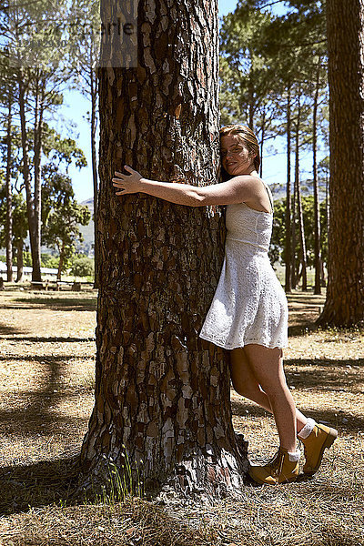 Lächelnde junge Frau umarmt einen Baum im Wald