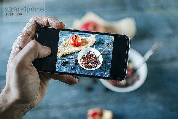 Männerhand beim Fotografieren mit Smartphone von Schale mit Dessert aus Mandeln  Granatapfel und Schokolade