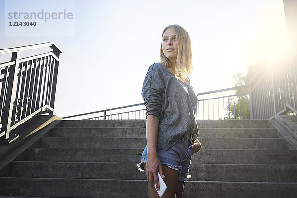Blonde junge Frau mit Smartphone im Gegenlicht vor der Treppe stehend
