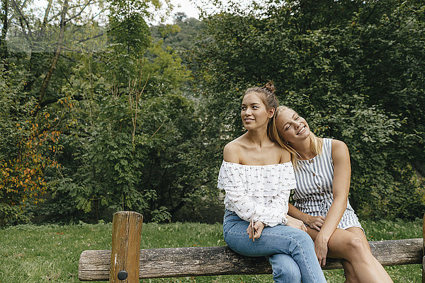 Zwei glückliche junge Frauen sitzen auf einem Zaun in einem Park.