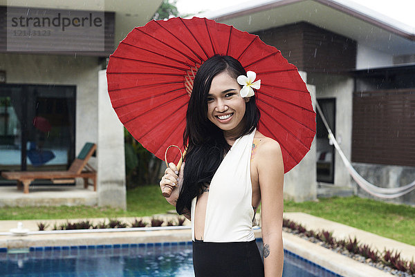 Porträt einer lächelnden Frau mit Blume im Haar  die einen roten Regenschirm an einem Swimmingpool hält.