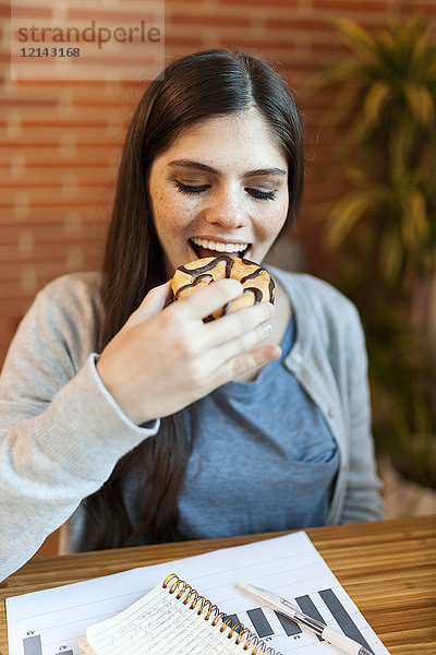Glückliche Geschäftsfrau beim Essen eines Donuts am Schreibtisch im Büro