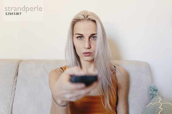 Junge Frau sitzt zu Hause auf der Couch mit Fernbedienung