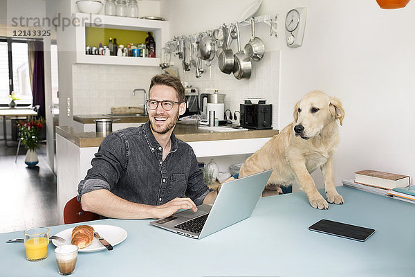 Porträt eines glücklichen Mannes mit Hund mit Laptop in der Küche zu Hause