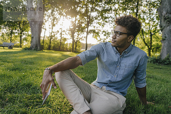 Junger Mann sitzt mit mobilem Gerät im Park und schaut sich um