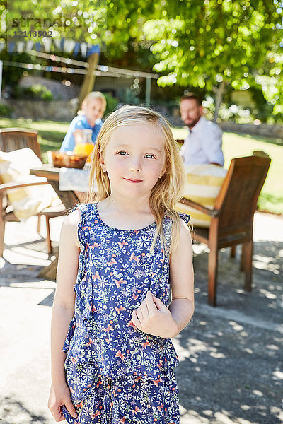 Porträt eines lächelnden Mädchens mit Eltern im Hintergrund am Gartentisch