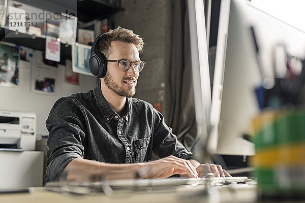 Lächelnder junger Mann mit Kopfhörern arbeitet am Computer zu Hause am Schreibtisch