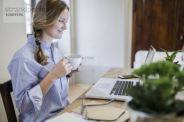 Lächelnde Frau am Schreibtisch mit Tasse Kaffee und Laptop