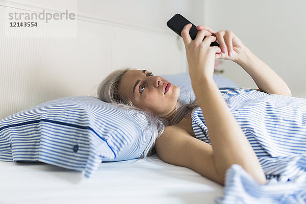 Seriöse junge Frau liegt im Bett und überprüft ihr Handy.