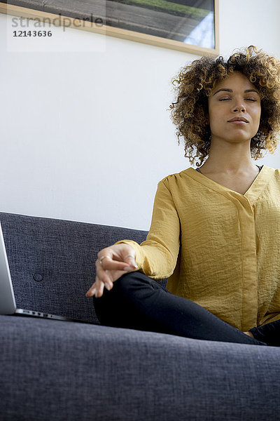 Junge Frau sitzt zu Hause auf der Couch neben dem Laptop und meditiert.