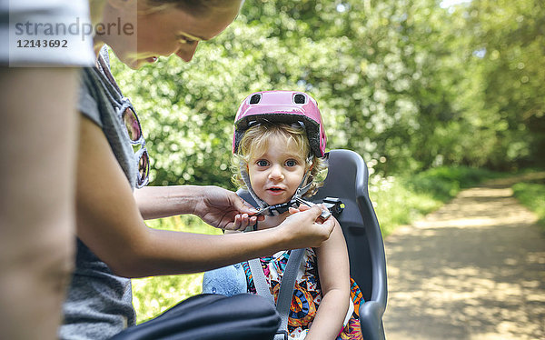 Kleines Mädchen sitzt auf einem Kindersitz für Fahrrad und ihre Mutter stellt ihren Helm ein