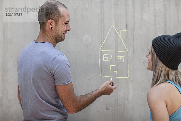 Mann und Frau zeichnen ein Haus mit Kreide auf Betonwand