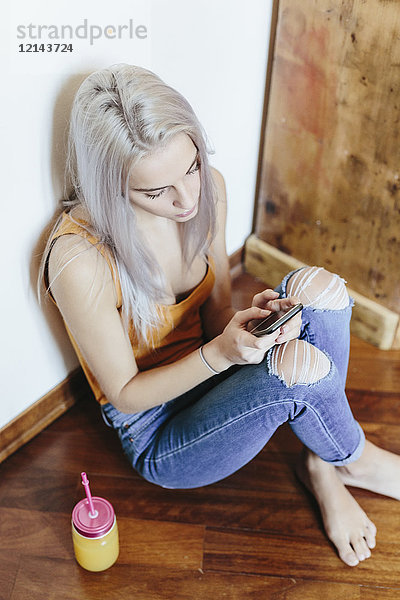 Junge Frau sitzt zu Hause auf dem Boden und benutzt ein Smartphone.