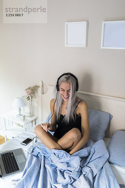 Glückliche junge Frau im Bett mit Handy und Kopfhörer