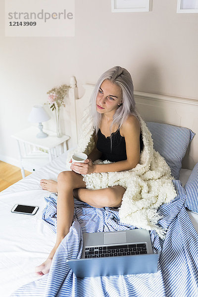 Nachdenkliche junge Frau im Bett mit Tasse Kaffee und Laptop