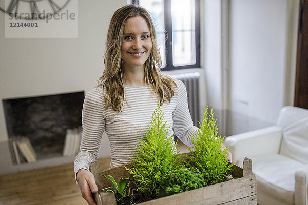 Porträt einer lächelnden Frau  die eine Kiste mit Pflanzen zu Hause trägt.