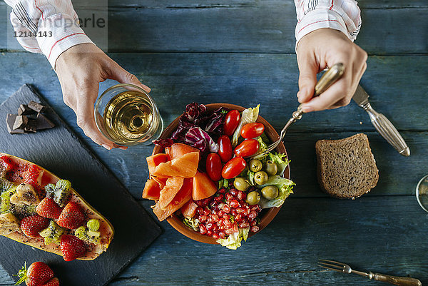 Nahaufnahme von Frauenhänden beim Essen von Salat mit Tomate  Granatapfel  Papaya und Oliven  Papaya mit Früchten und Weinglas