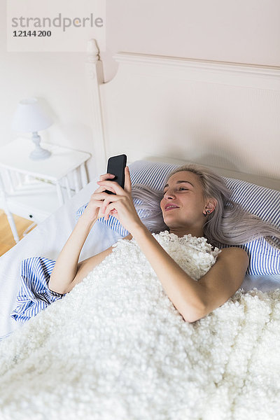 Lächelnde junge Frau  die im Bett liegt und ihr Handy überprüft.