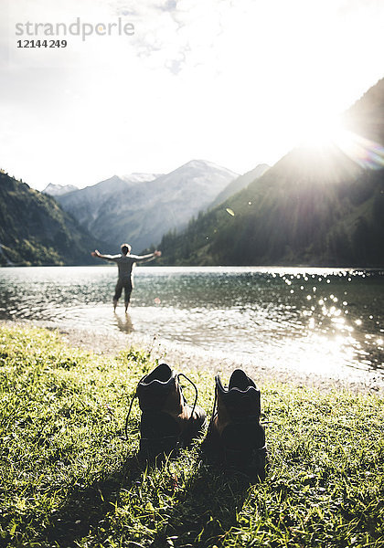 Österreich  Tirol  Wanderschuhe und Mann mit ausgestreckten Armen im Bergsee