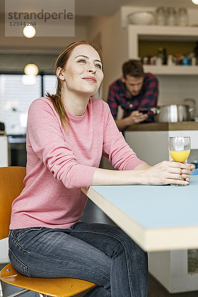 Lächelnde junge Frau mit Glas Orangensaft in der Küche zu Hause mit Mann im Hintergrund